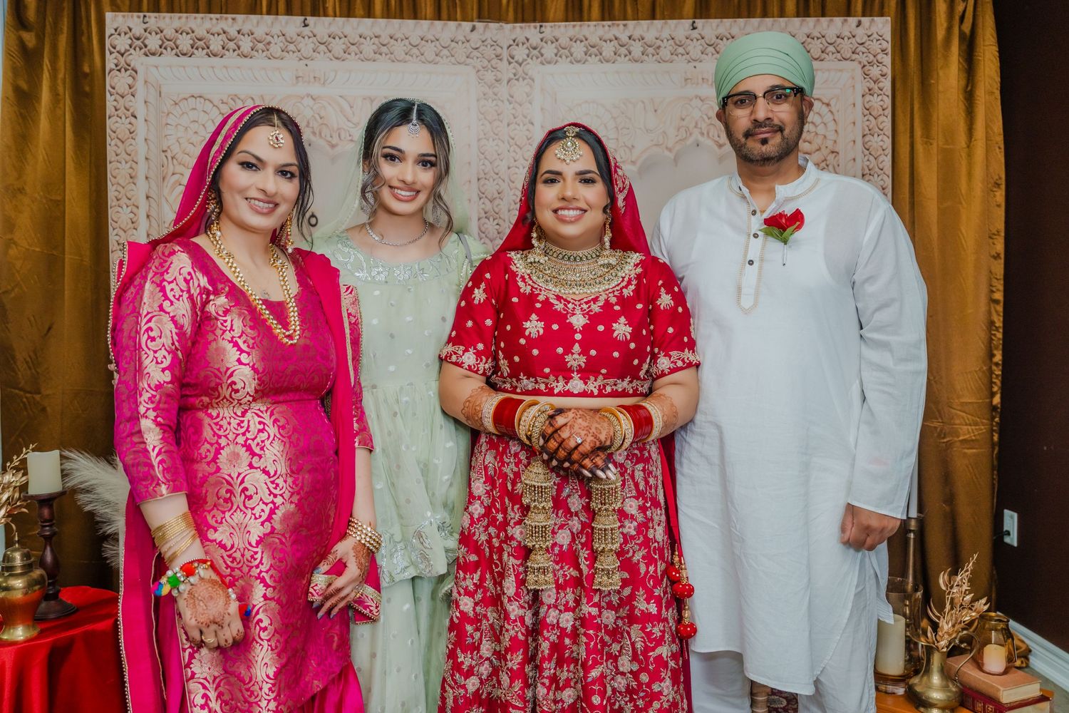Four people dressed in traditional South Asian wedding attire pose together against an ornate patterned backdrop.