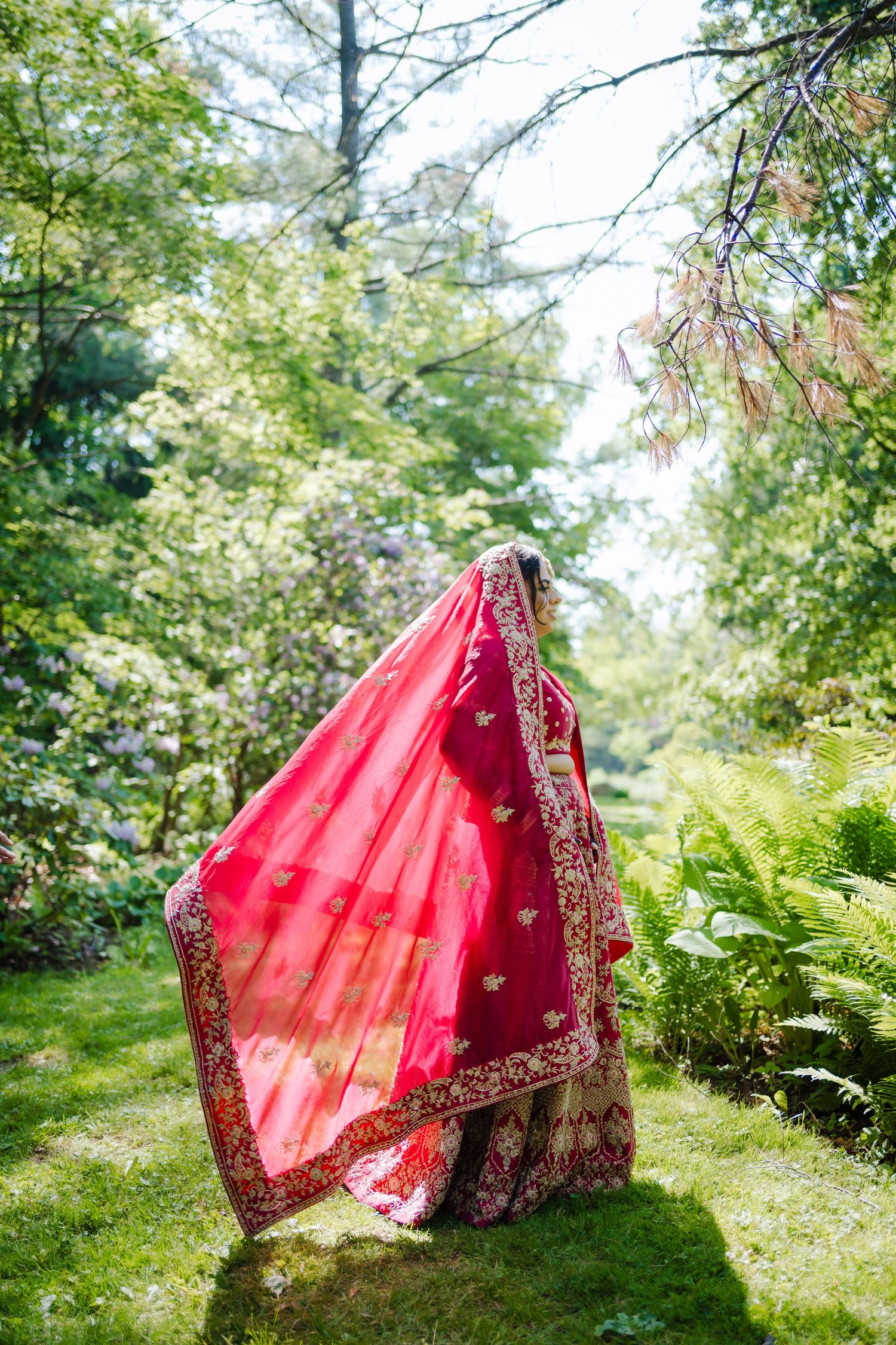 A vibrant red and gold embroidered sari flows elegantly in a lush garden setting surrounded by green foliage.