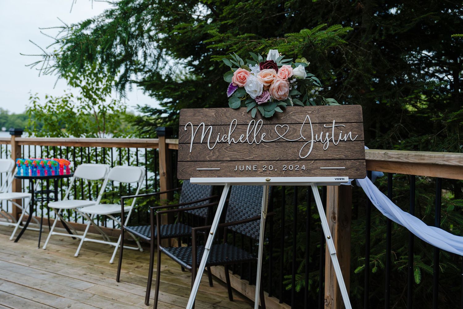Rustic wooden wedding welcome sign with floral decor displayed on an outdoor deck railing.