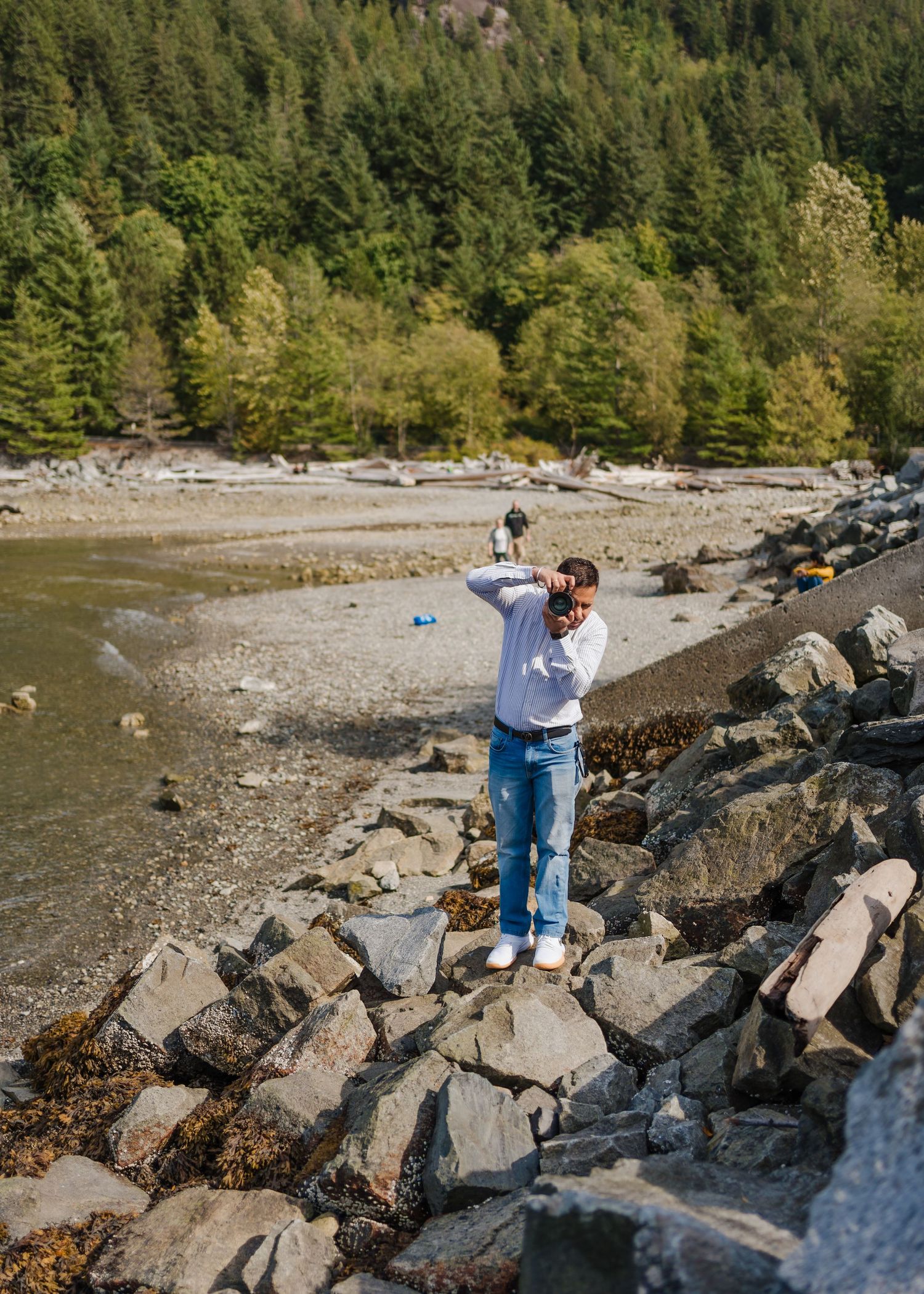 Person stands on rocky shoreline overlooking a serene lake surrounded by forest.