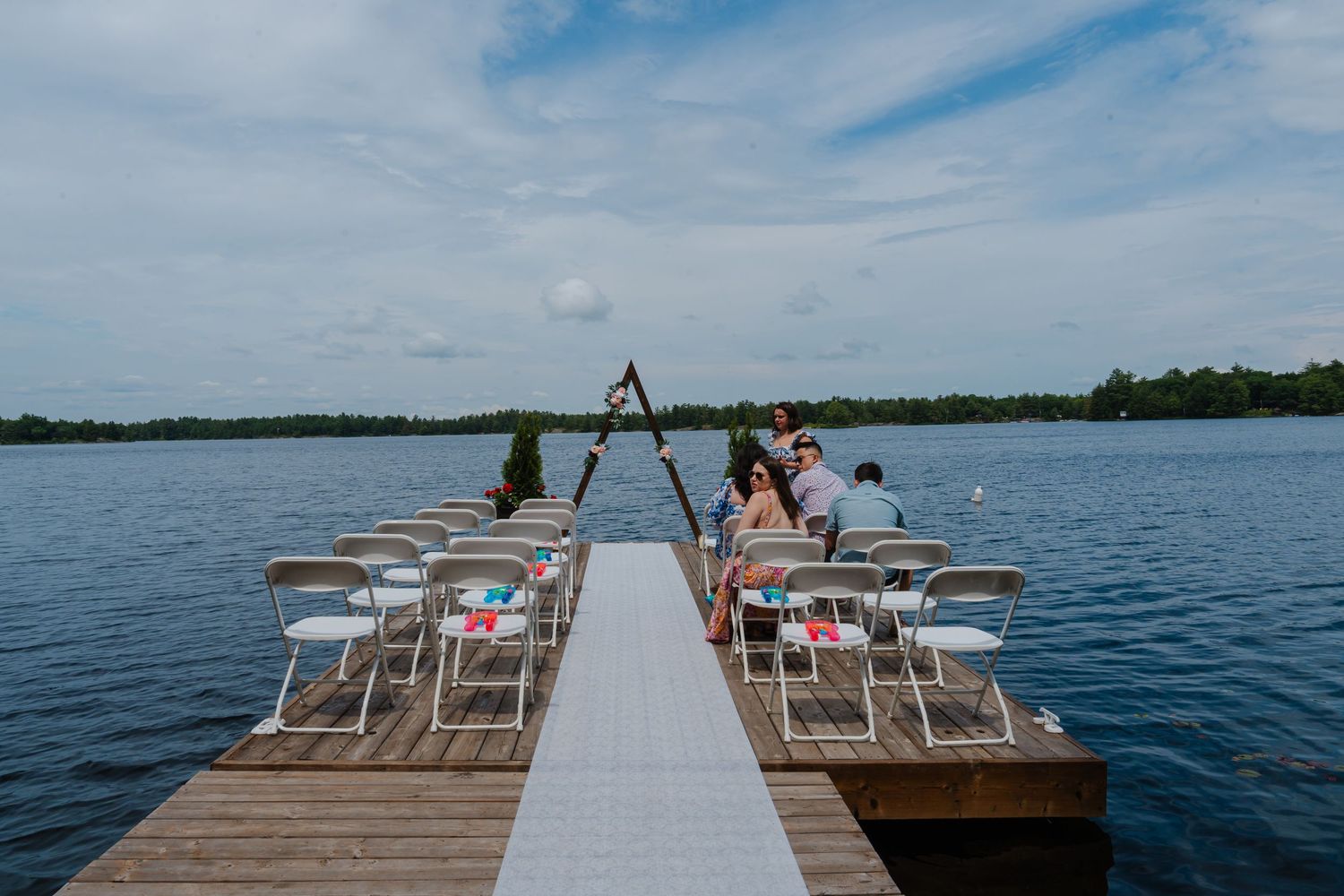 White chairs line wooden dock for waterfront wedding ceremony under blue sky.