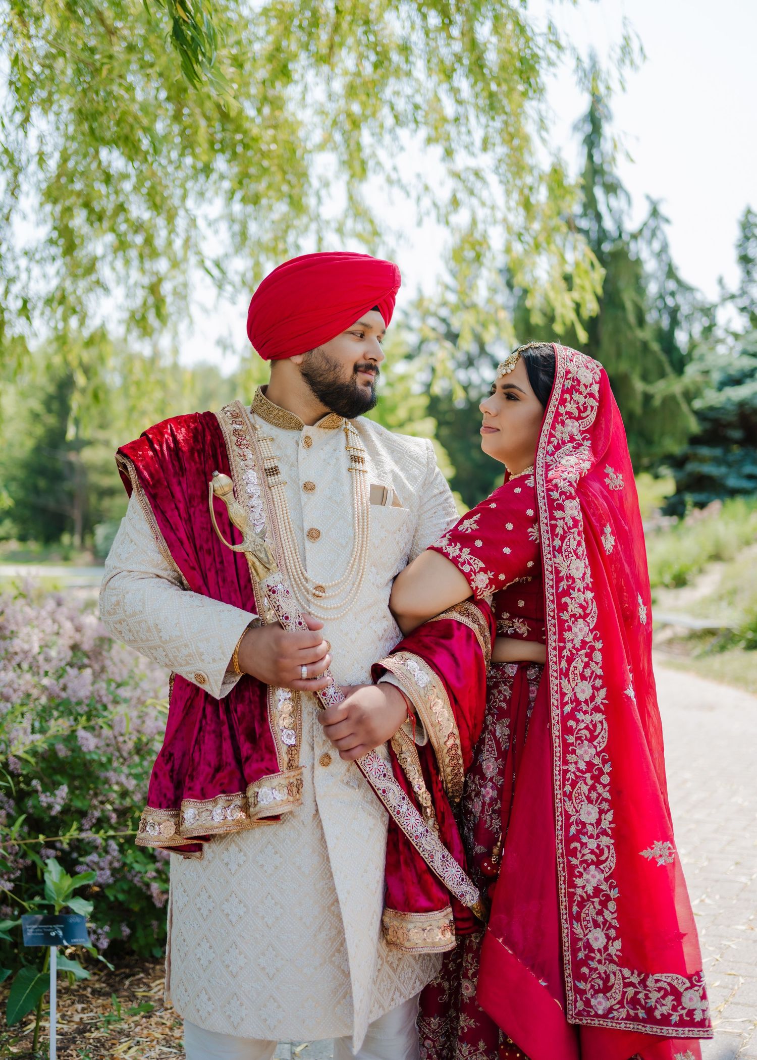 Traditional Sikh wedding couple in elegant red and white attire pose lovingly in a garden setting with willow trees.