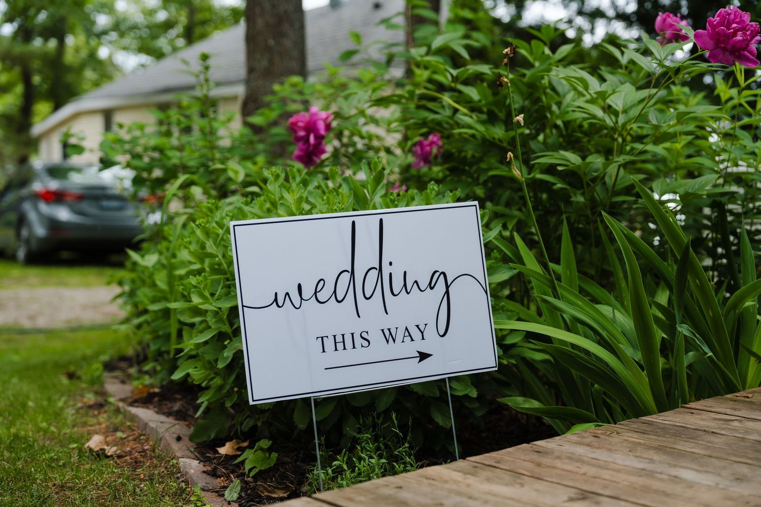 A rustic wedding directional sign placed in a garden with pink flowers and greenery.