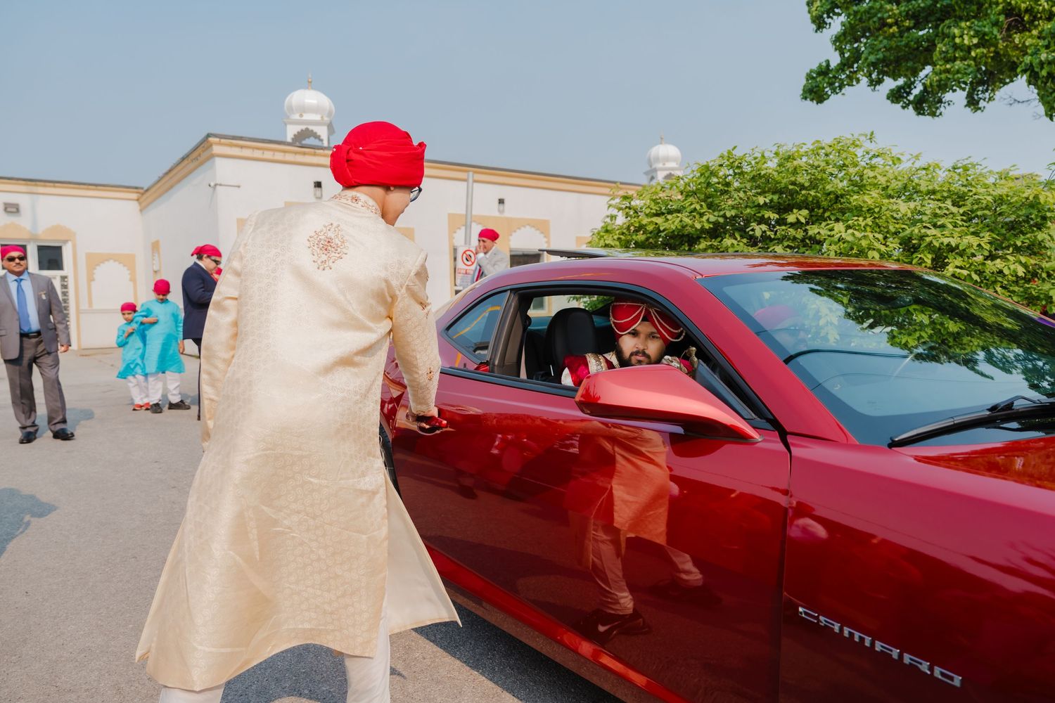 A person wearing formal beige attire and red headwear interacts with someone in a red car outside a building.