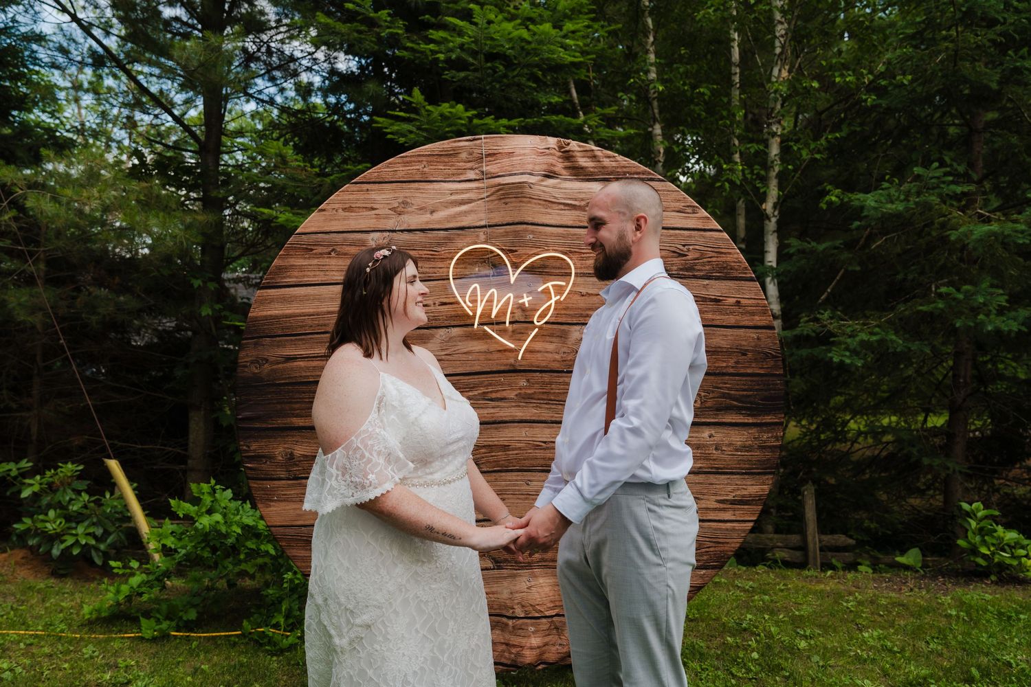 A couple stands together in front of a round wooden backdrop with a heart design at an outdoor woodland wedding ceremony.