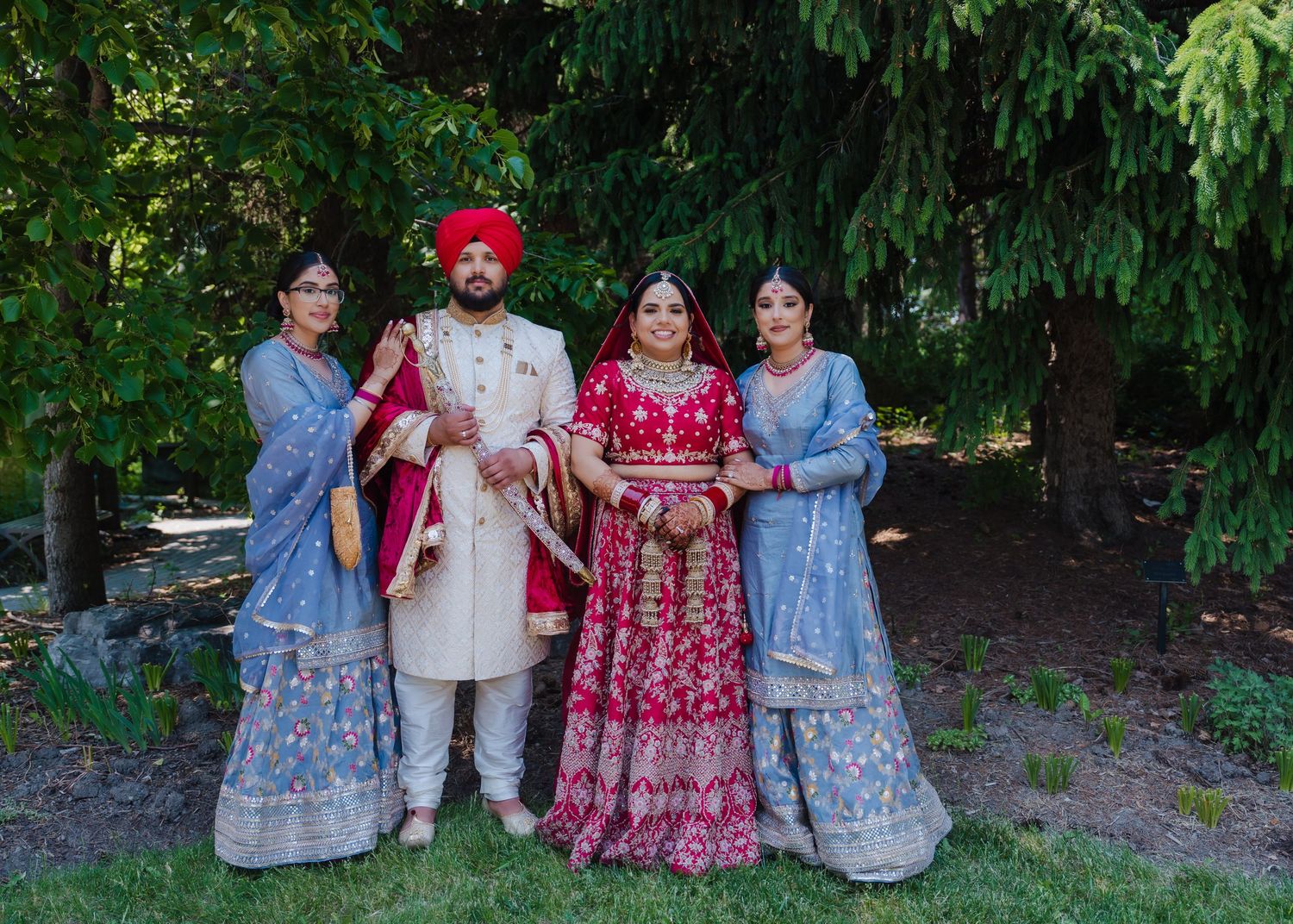 Wedding party in traditional Indian attire posing outdoors with bride in red and bridesmaids in blue lehengas.