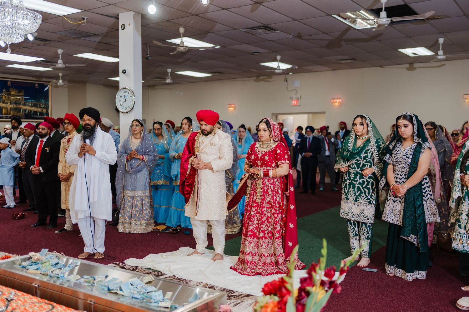 Attendees in traditional South Asian attire gather for prayer during a religious ceremony in a community space.