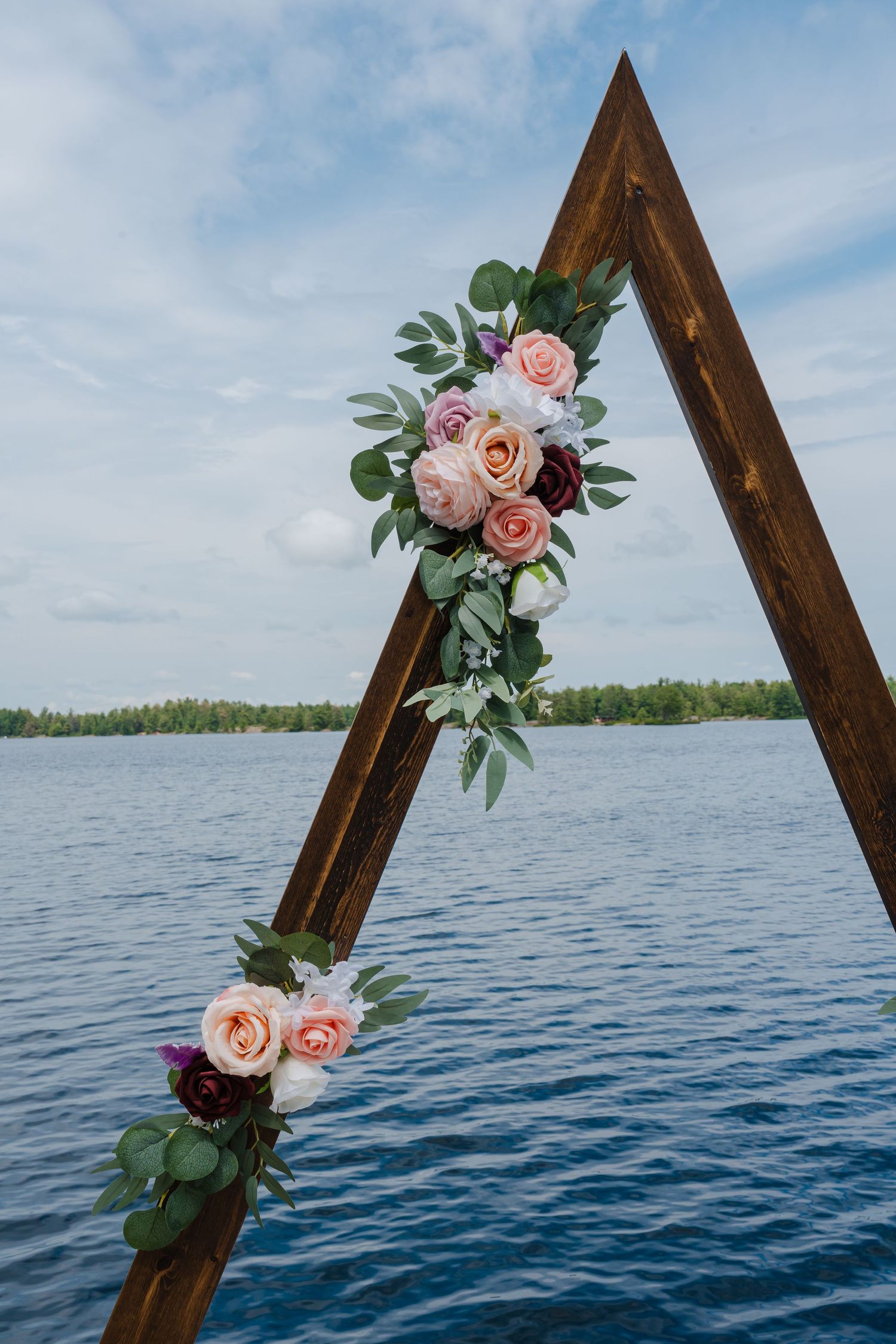 A wooden triangle arch decorated with pink roses and greenery stands against a serene lake backdrop with blue skies.
