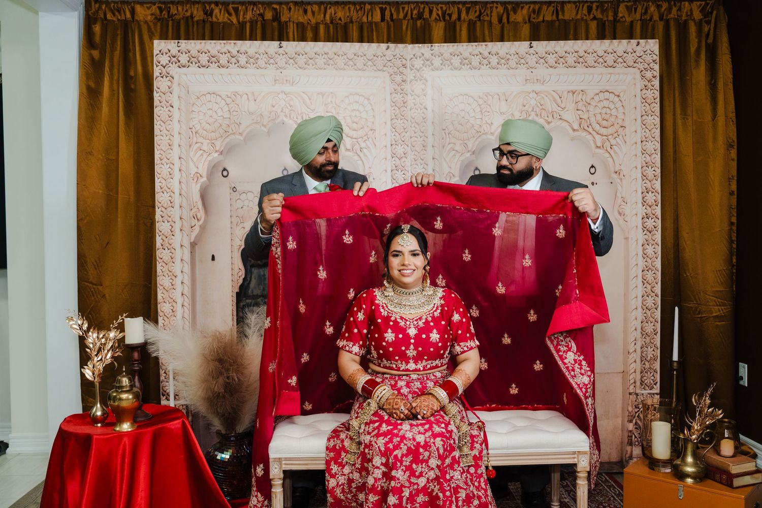 A bride in red traditional attire sits while two men in turbans hold a red fabric backdrop during a ceremony.