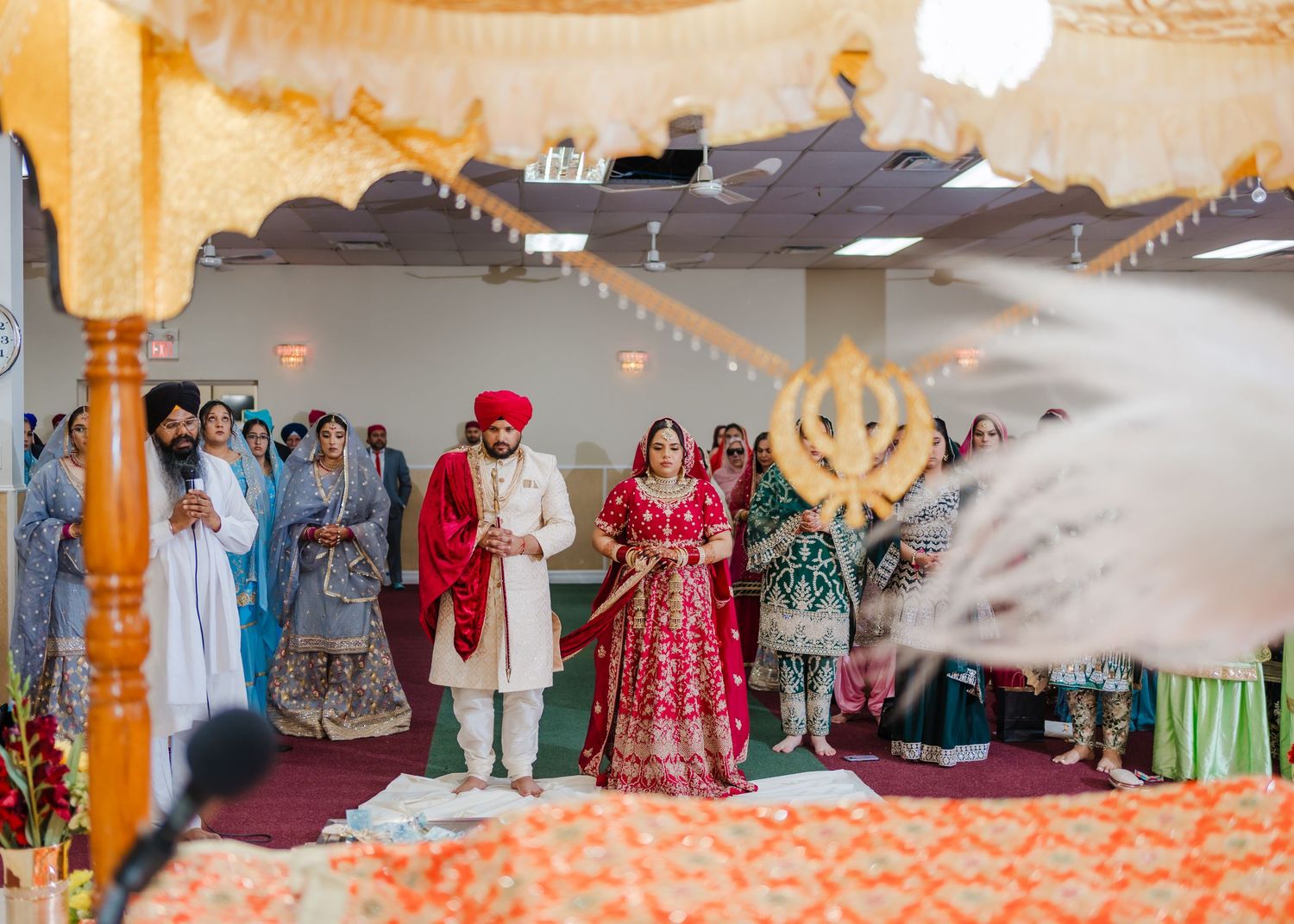 Ornate Sikh wedding ceremony with decorative orange drapery and Khanda symbol visible in background.