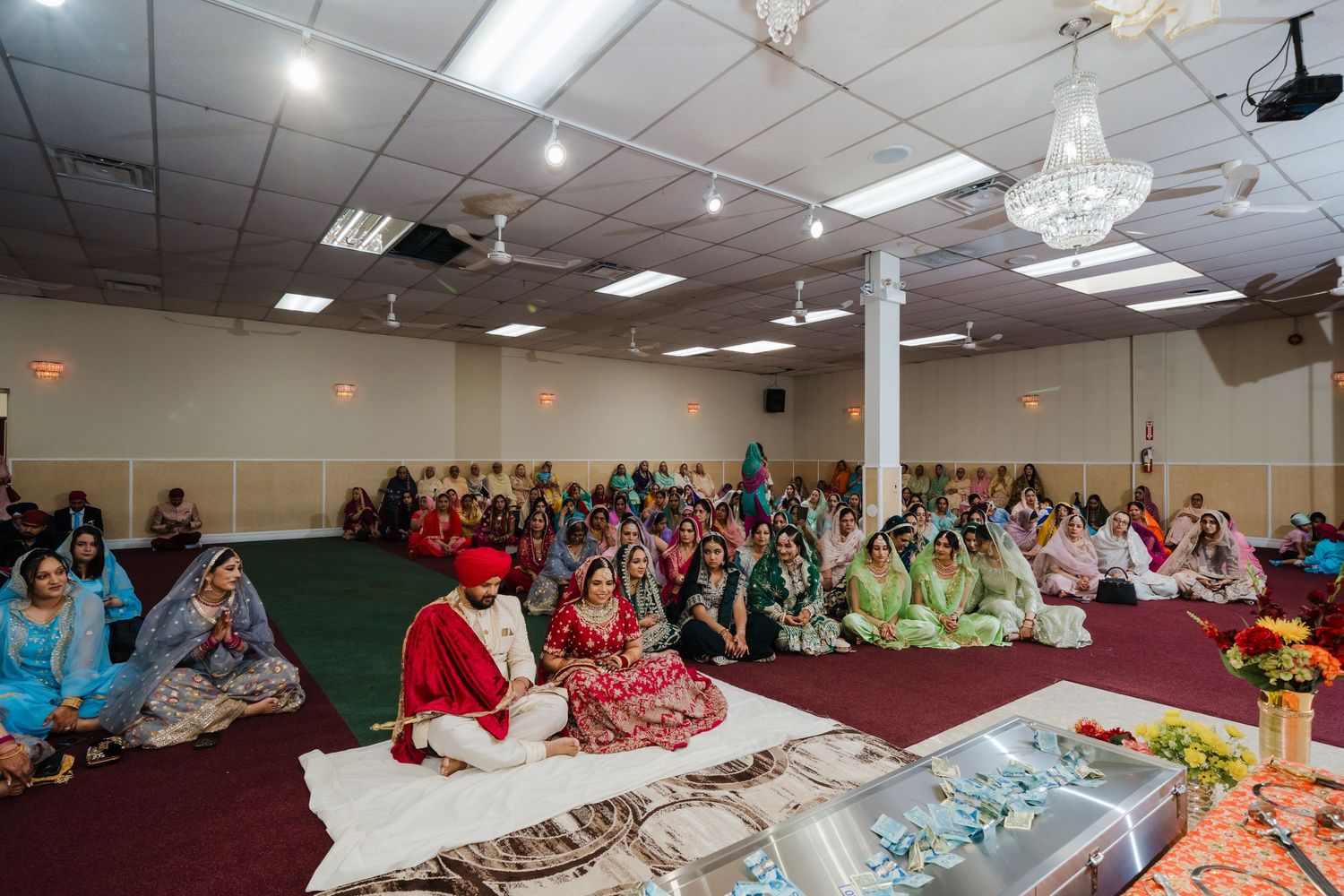 Large group gathered in a community hall for a Sikh wedding ceremony with traditional decorations.