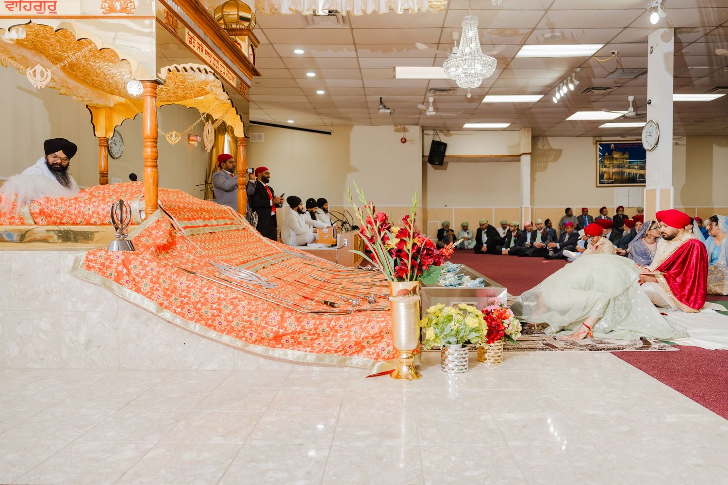 Traditional Indian wedding ceremony in progress with ornate orange and gold decorations in a bright indoor venue.