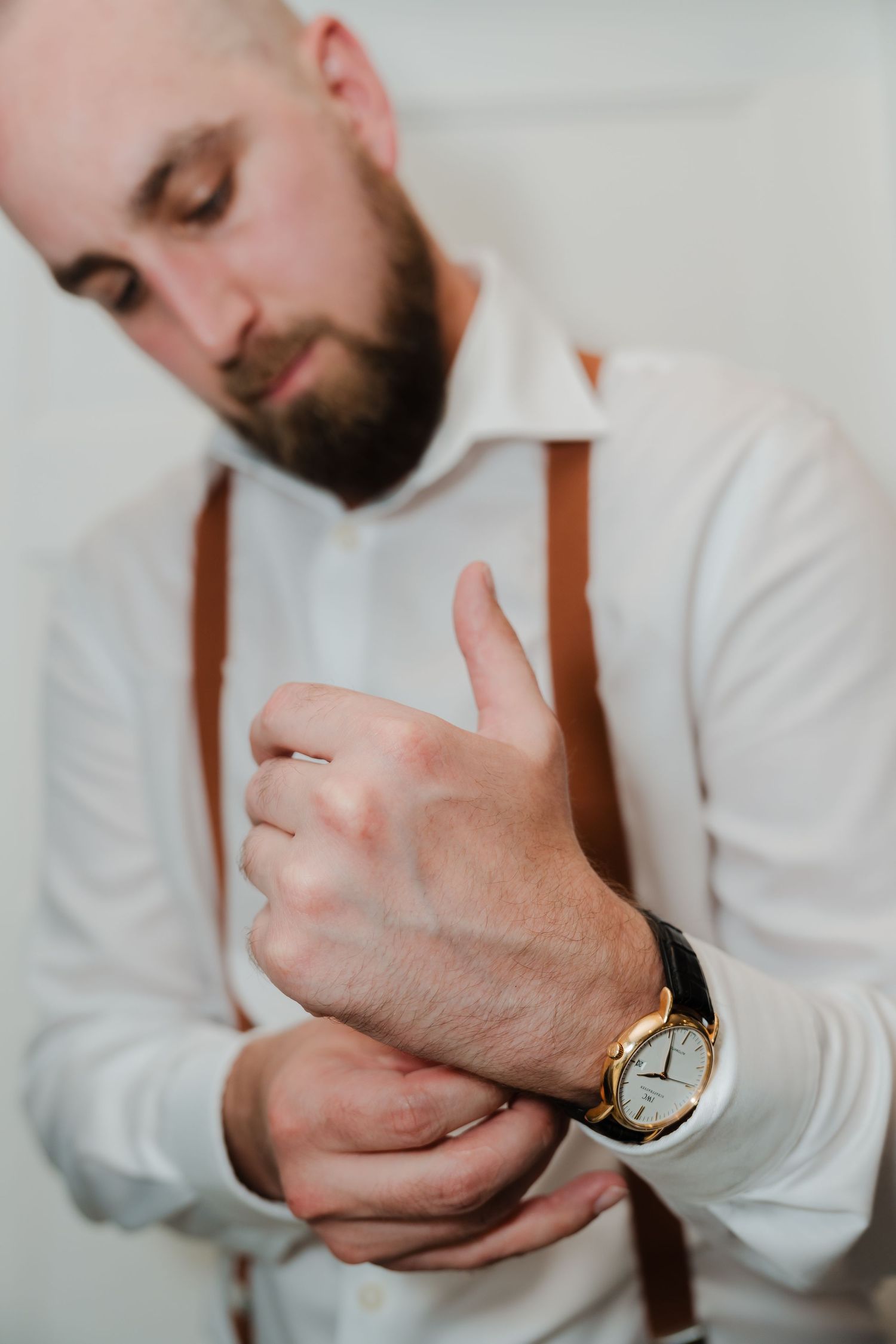 Close-up shot of groom adjusting white dress shirt cuff while wearing brown suspenders and gold watch.