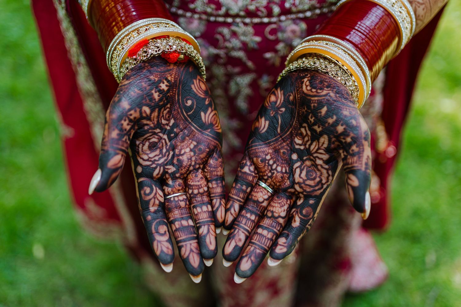 Intricate henna designs adorning hands wearing traditional gold bangles against a red dress and green grass background.
