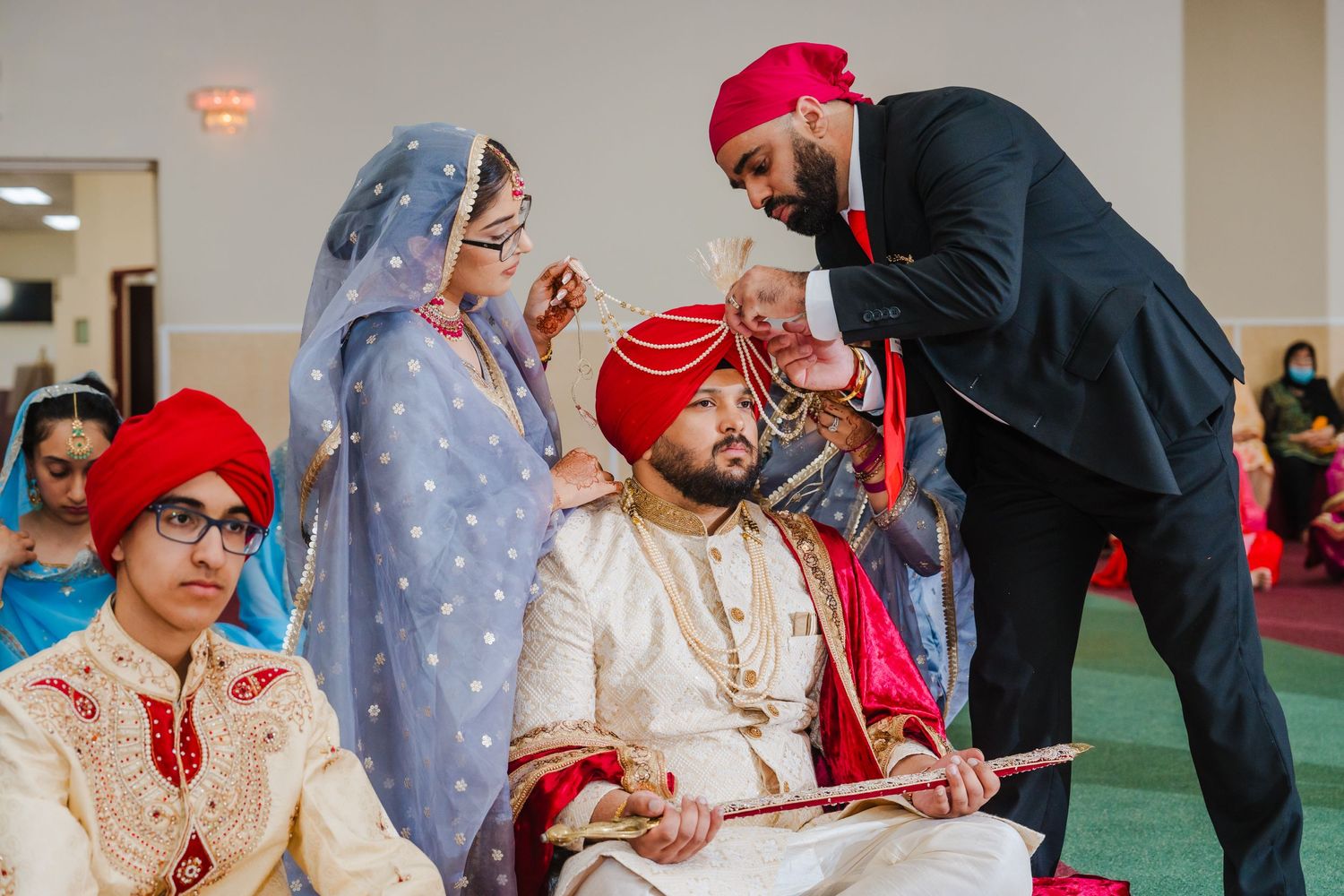 People in traditional Sikh attire gather for a cultural wedding ceremony on a carpeted floor indoors.