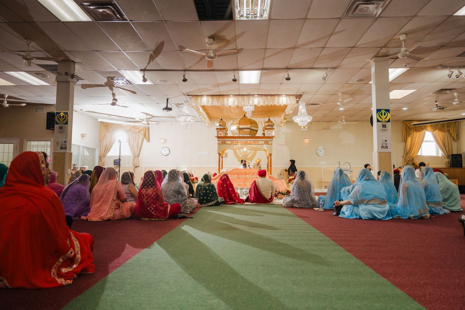 A group of worshippers in colorful traditional clothing sits together on a carpeted floor in a well-lit prayer space.