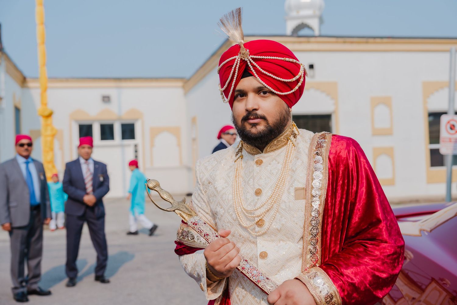 Groom in traditional red and gold Sikh wedding attire with turban holding ceremonial items.