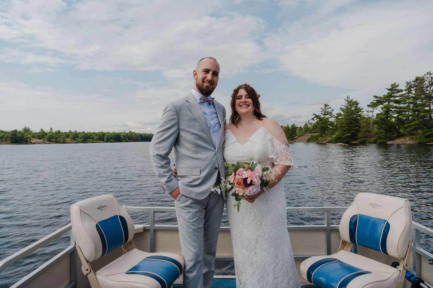 Wedding couple stands together on a boat deck with a scenic lake and pine trees in the background.