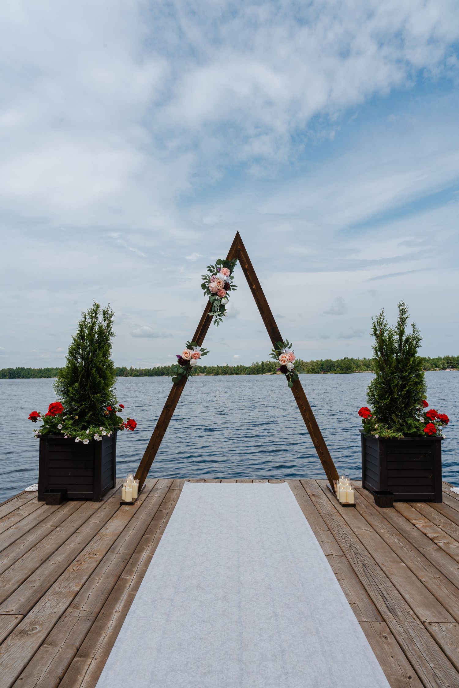 A wooden triangular wedding arch decorated with flowers and greenery stands on a dock overlooking a serene lake.