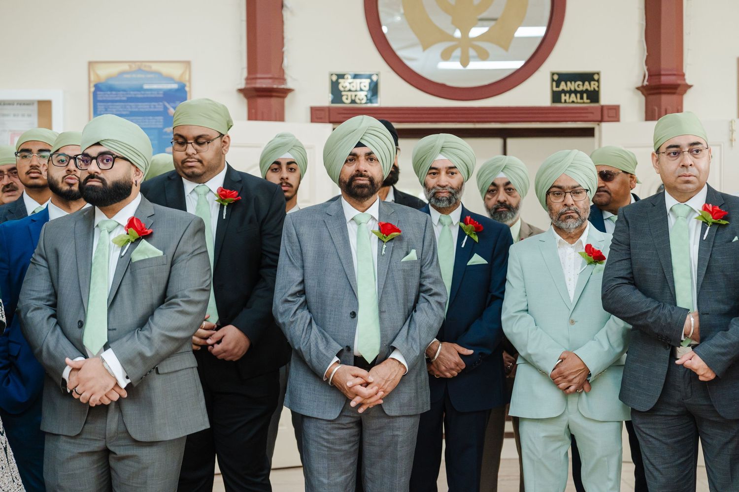 Group of men in formal grey suits with red boutonnieres standing in a row at Sikh wedding ceremony.