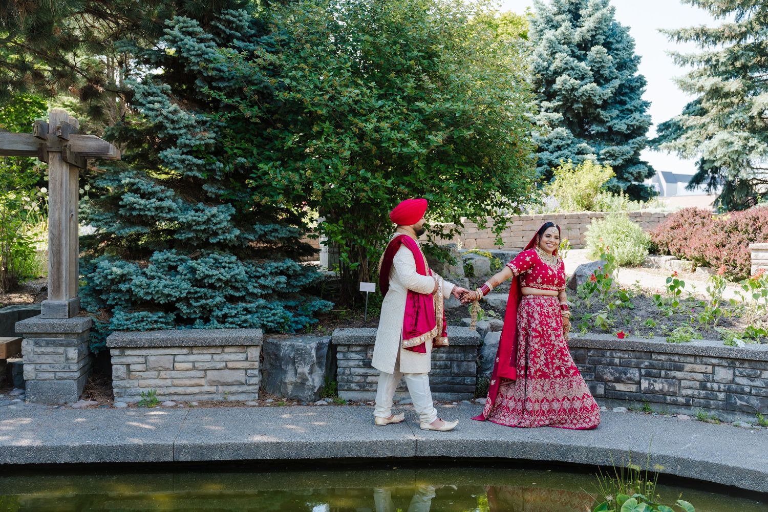 A couple in traditional Indian wedding attire stands on a stone bridge near evergreen trees and a pond.