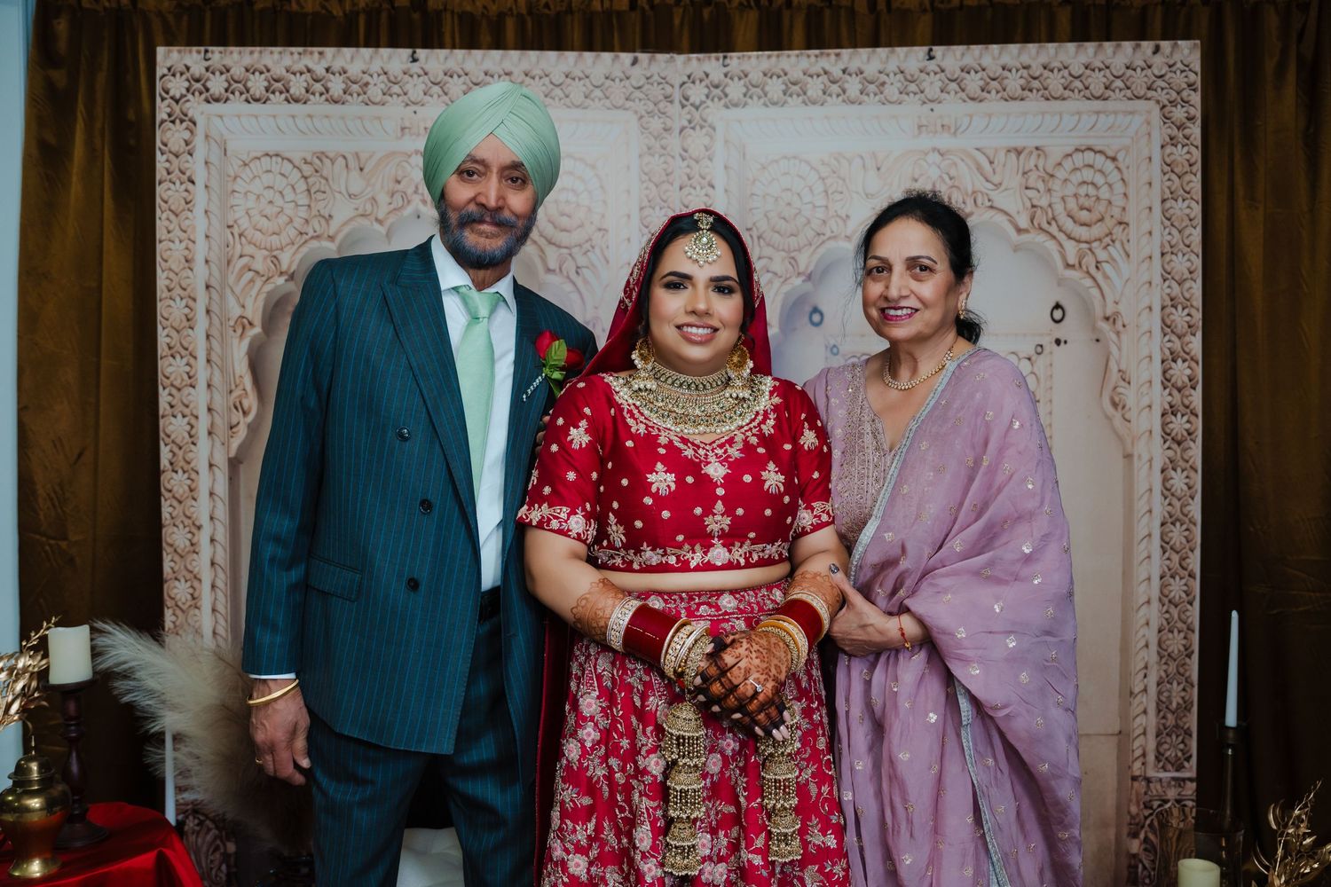Three people wearing traditional Indian wedding attire pose against a decorated backdrop.