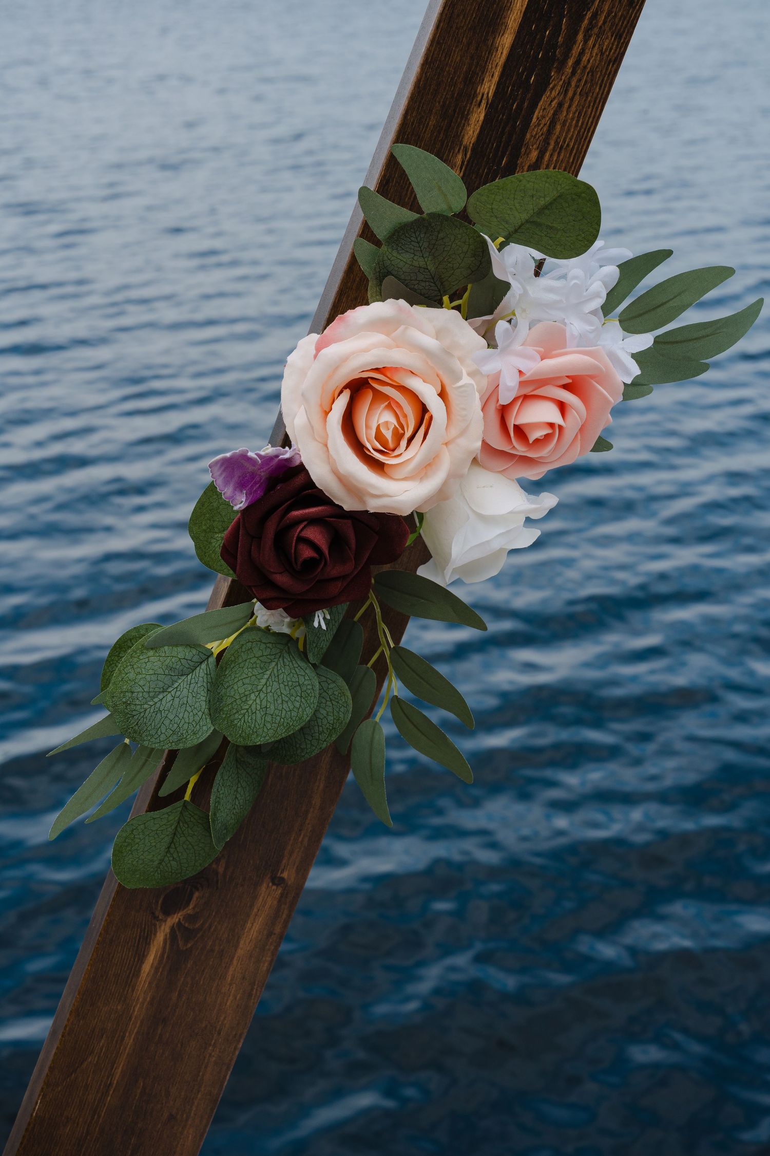 Pink and burgundy roses with eucalyptus leaves arranged on a wooden beam against ocean waters.