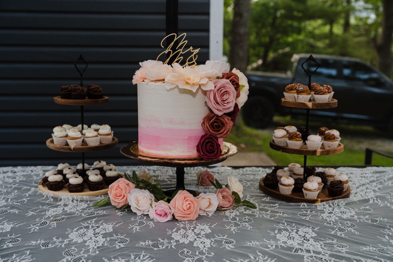 A pink ombre wedding cake decorated with roses sits on a lace tablecloth surrounded by chocolate cupcakes.