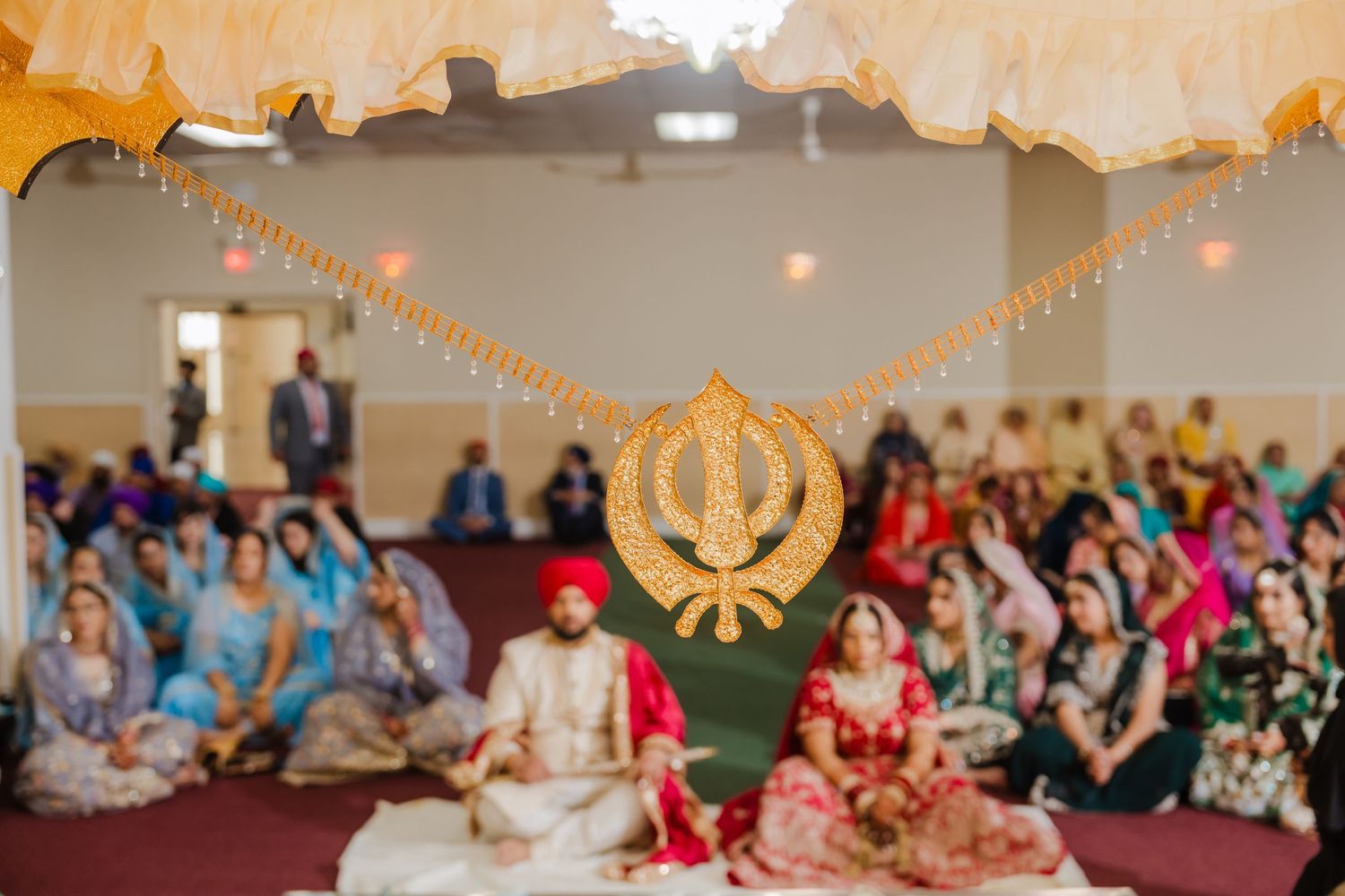 A Sikh wedding ceremony in a temple with the Khanda symbol hanging above and guests sitting on the floor.