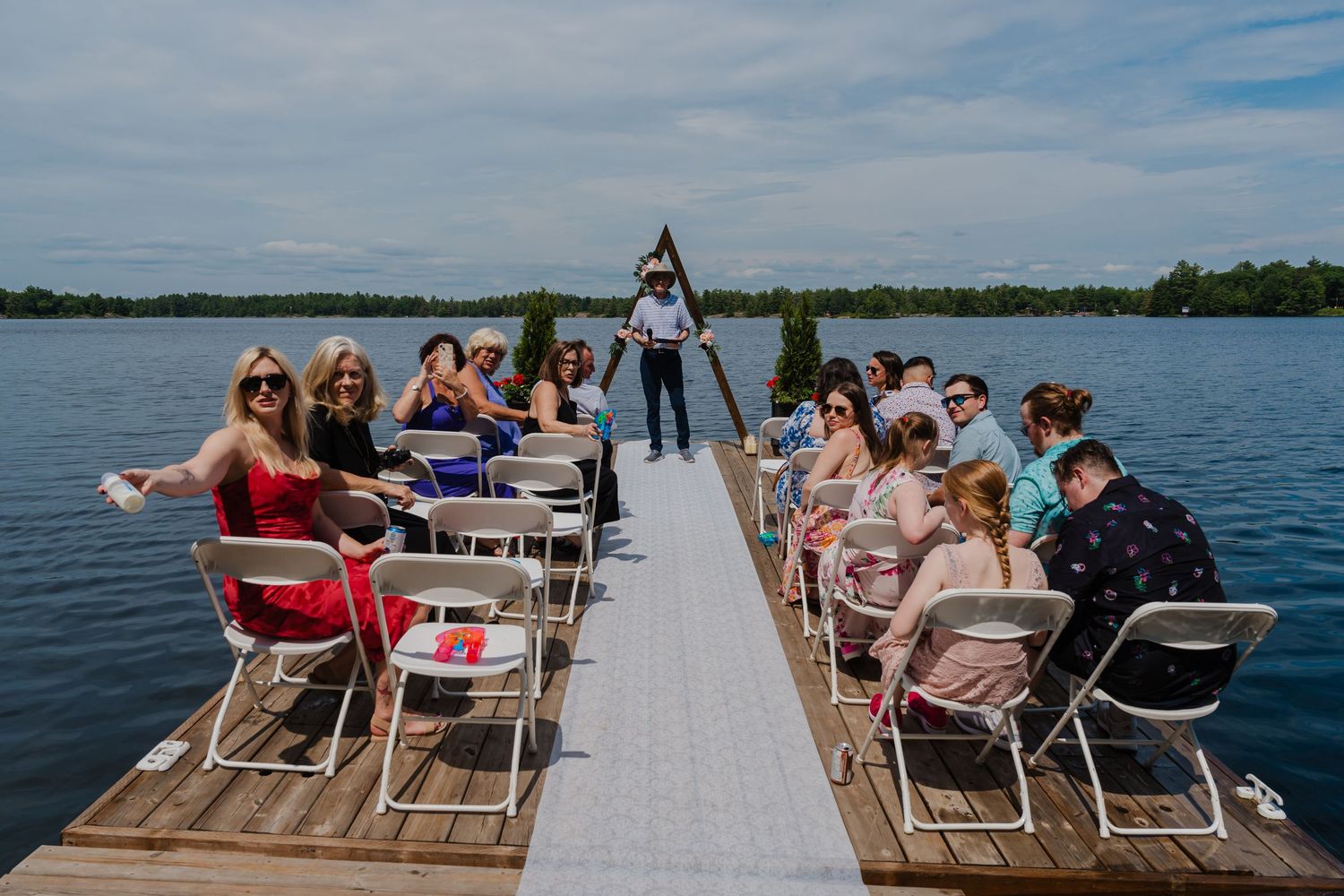Wedding guests seated on folding chairs along a wooden dock during a lakeside ceremony.