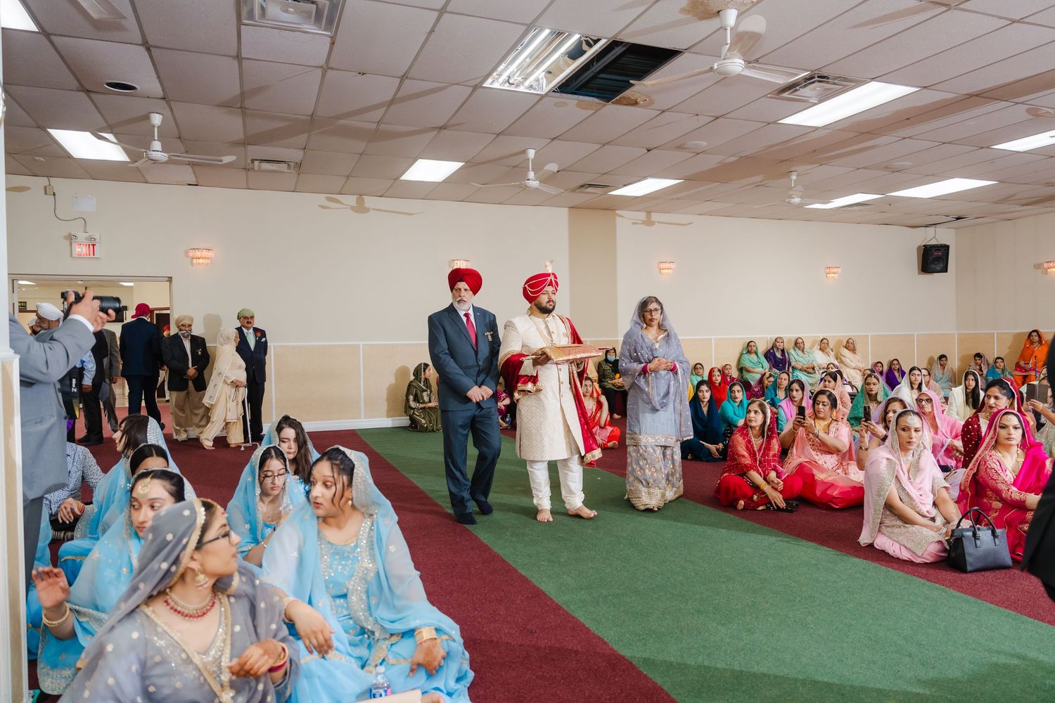 Wedding ceremony procession on green carpet with guests in traditional Indian formal wear seated on both sides.