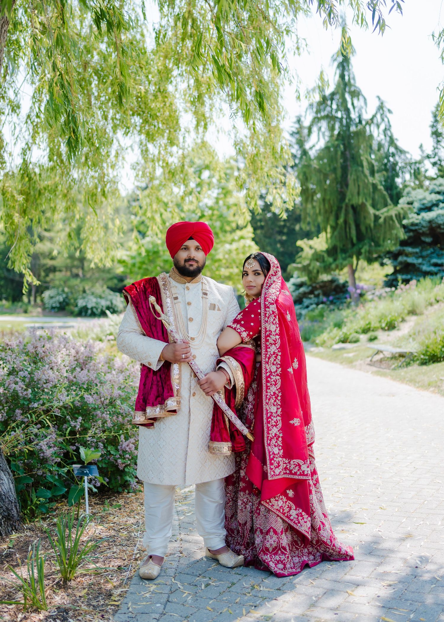 A couple in red and cream traditional wedding outfits pose on a garden path under weeping willows.