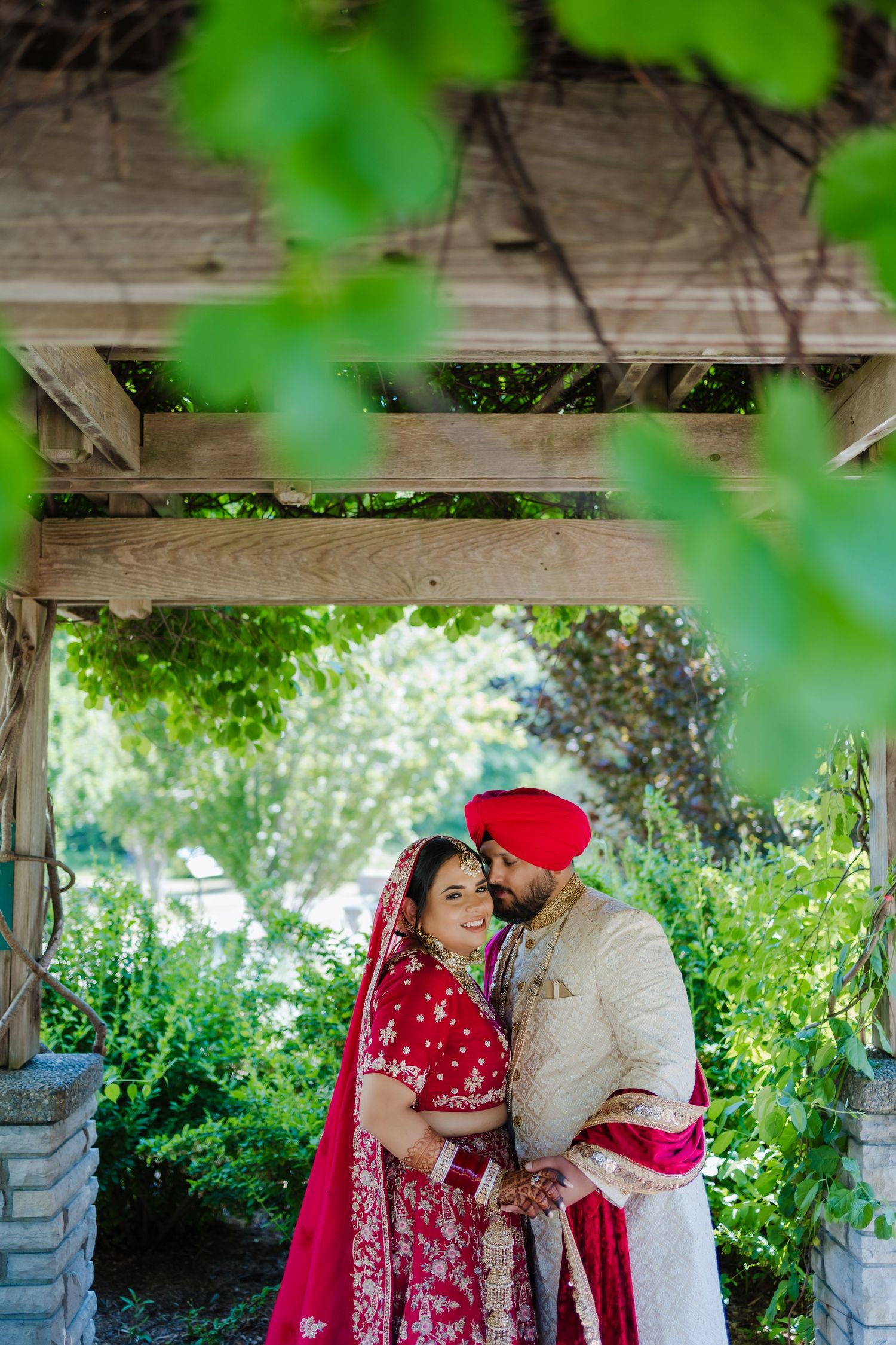 A couple in red and cream wedding attire embraces under a wooden pergola in a garden.