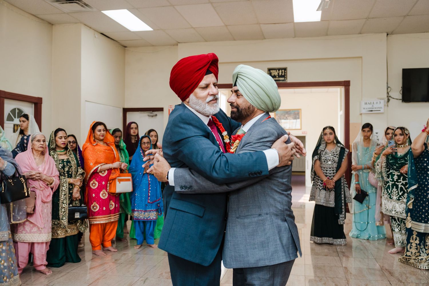 Two people wearing turbans embrace in a hallway while guests in colorful traditional attire watch in the background.
