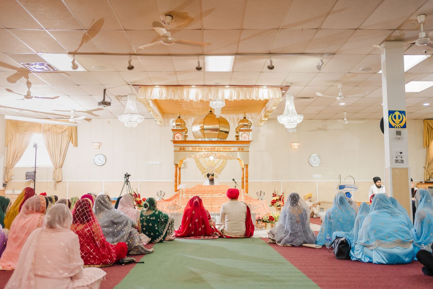 A group of worshippers in colorful traditional attire gather on green carpet in front of ornate golden decor during prayer.