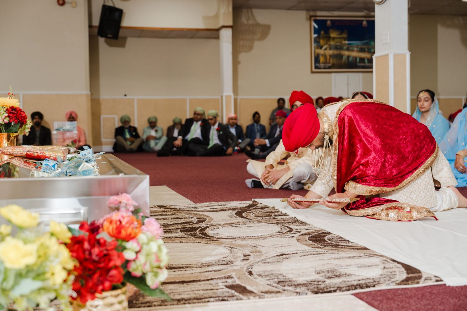 People gather in a mosque for prayer with a carpet adorned with intricate patterns and flowers in the foreground.