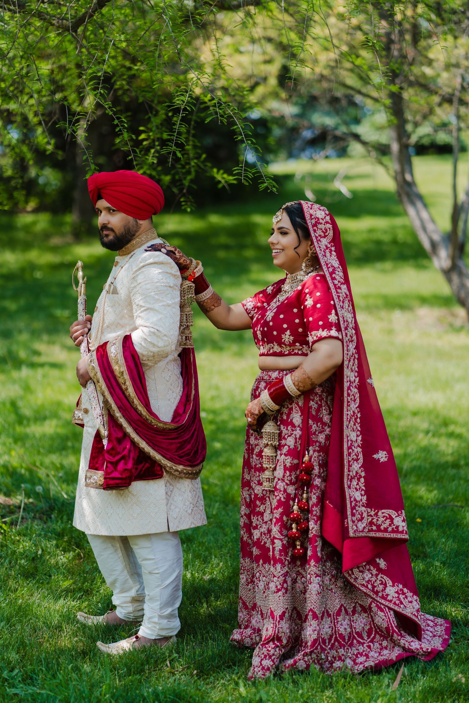 Traditional Sikh wedding couple in cultural attire pose together in a garden setting with green trees.