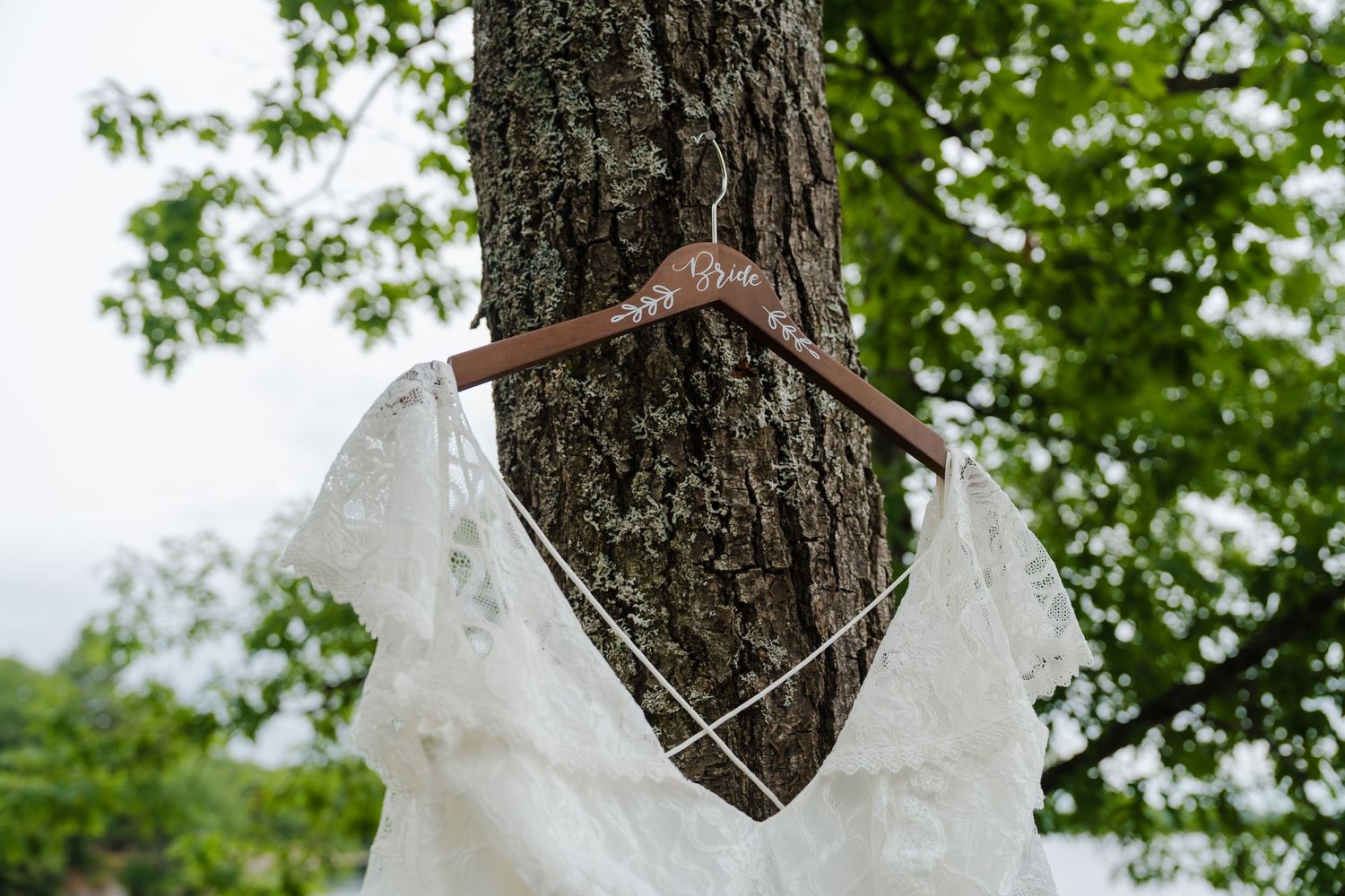 A white lace wedding dress hangs on a wooden hanger against a tree trunk with green foliage in the background.