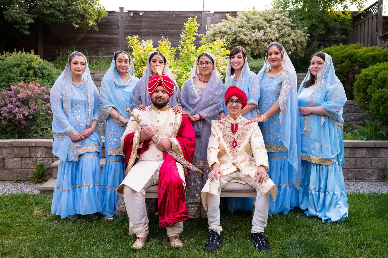 A wedding party in coordinated light blue and red traditional South Asian attire poses together on a garden lawn.
