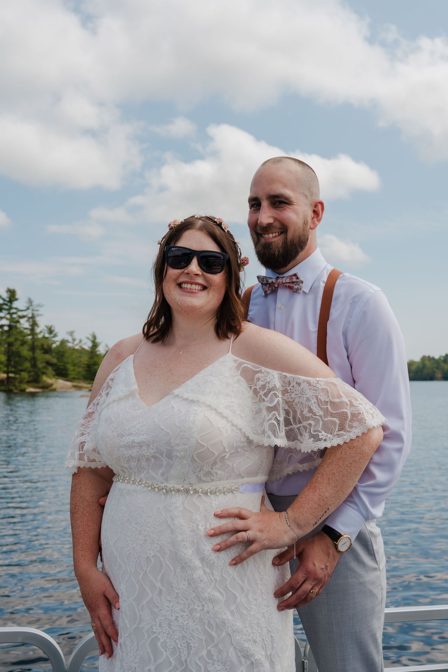 A couple poses together on a boat dock with a lake and trees in the background on their wedding day.