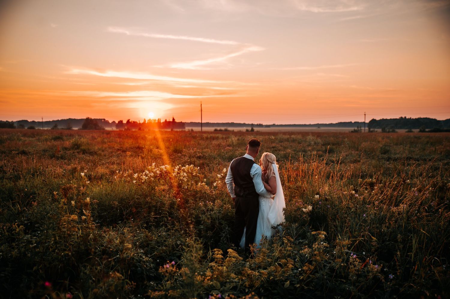 A couple stands in a golden meadow at sunset with warm orange light illuminating the wildflowers and landscape.