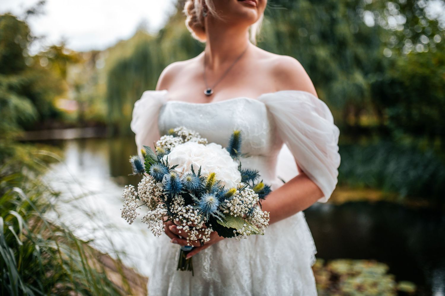A bridal bouquet with blue and white flowers held against a blurred outdoor background with water and trees.