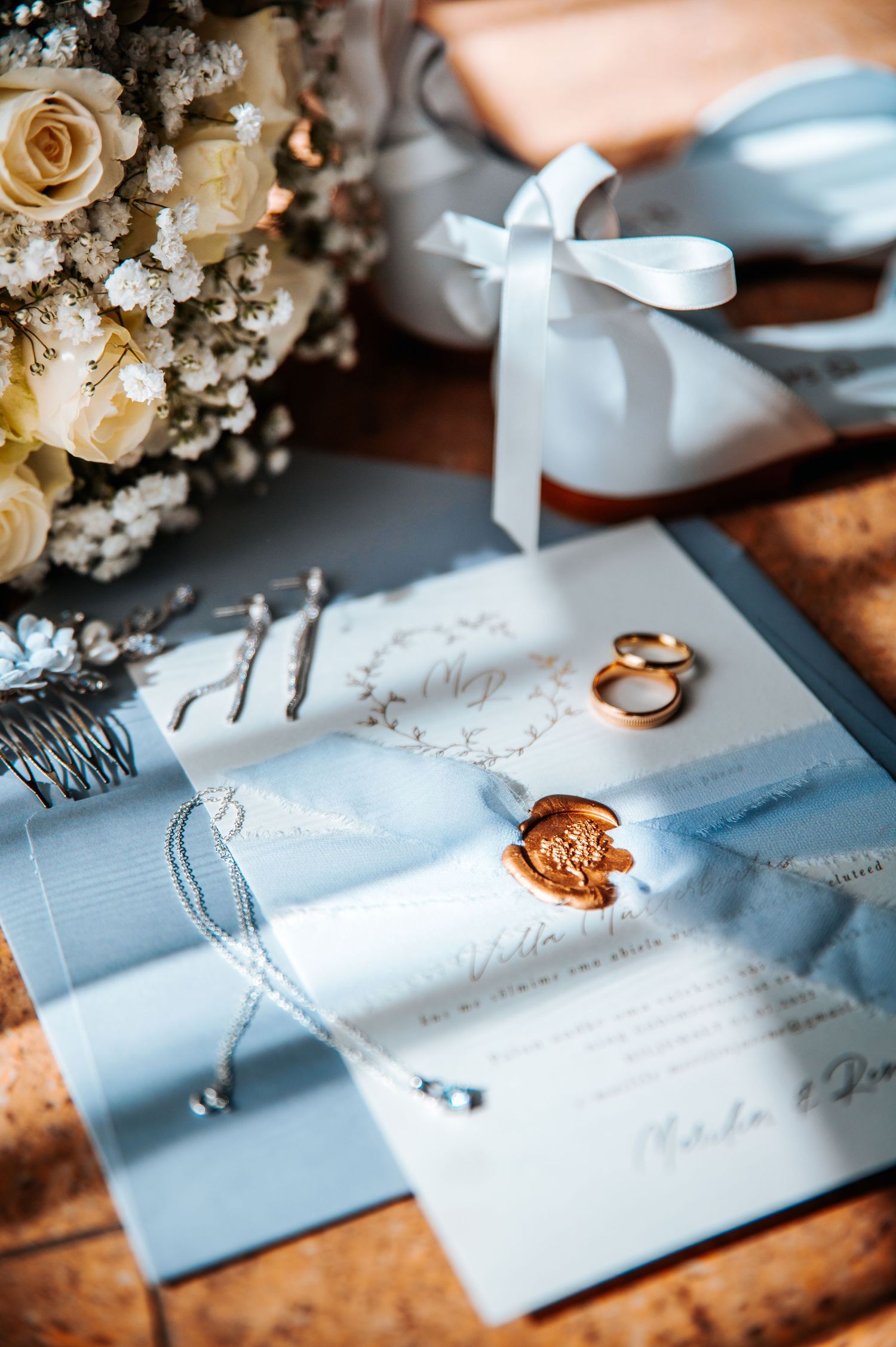 Wedding rings and invitation details styled with delicate flowers and ribbons on a light blue surface.