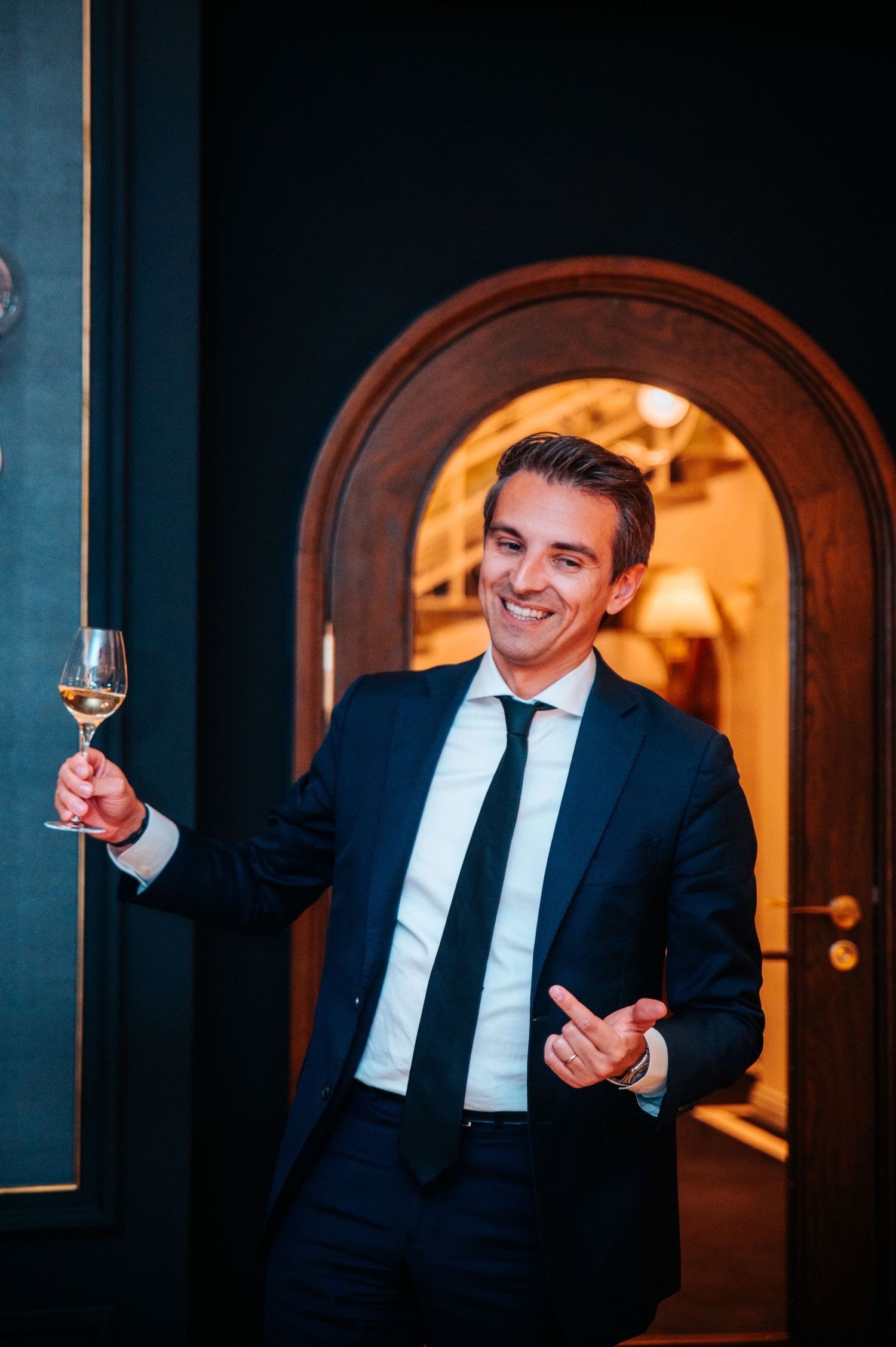 Person in formal attire enjoying wine in dimly lit elegant venue with arched doorways.