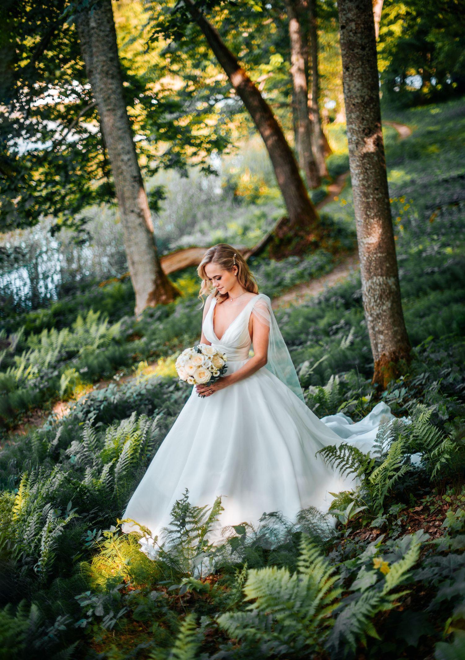 An elegant white wedding dress photographed in a lush forest setting with ferns and dappled sunlight.