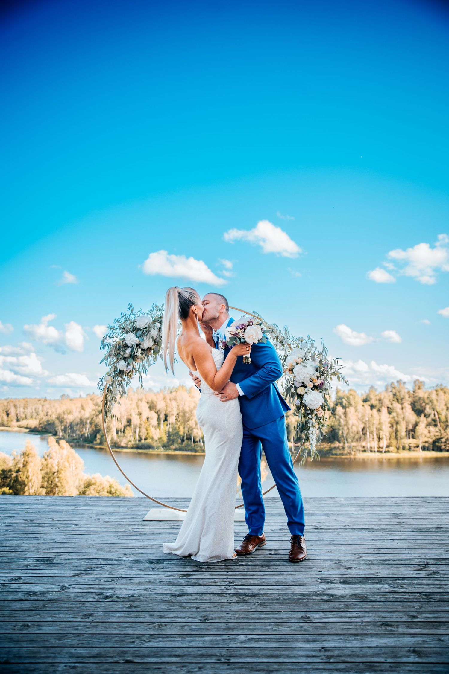 A couple in wedding attire shares a romantic dip pose on a wooden dock by a serene lake with autumn trees in the background.