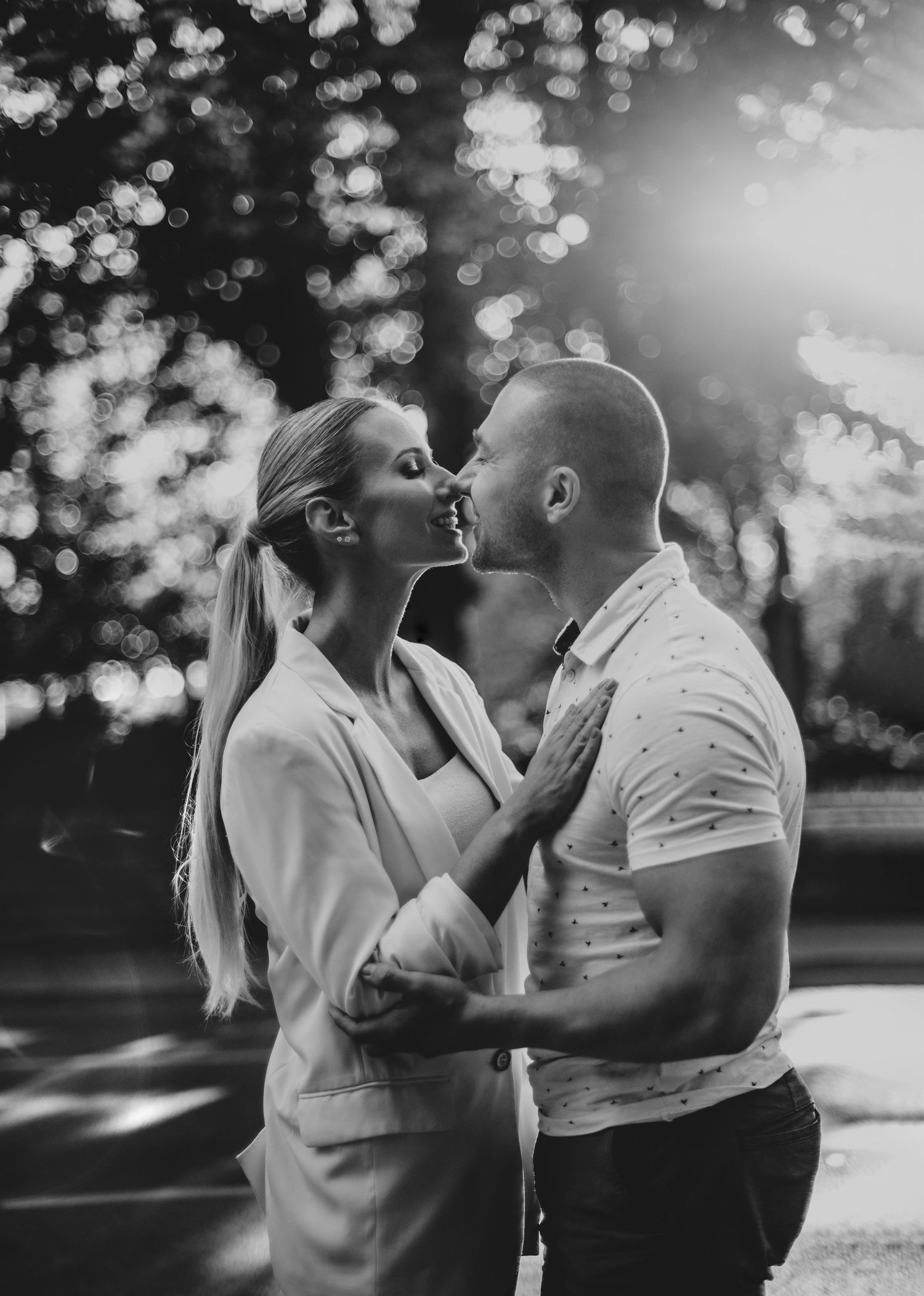 Black and white romantic couple sharing playful moments in a sunlit outdoor setting with bokeh effect in background.