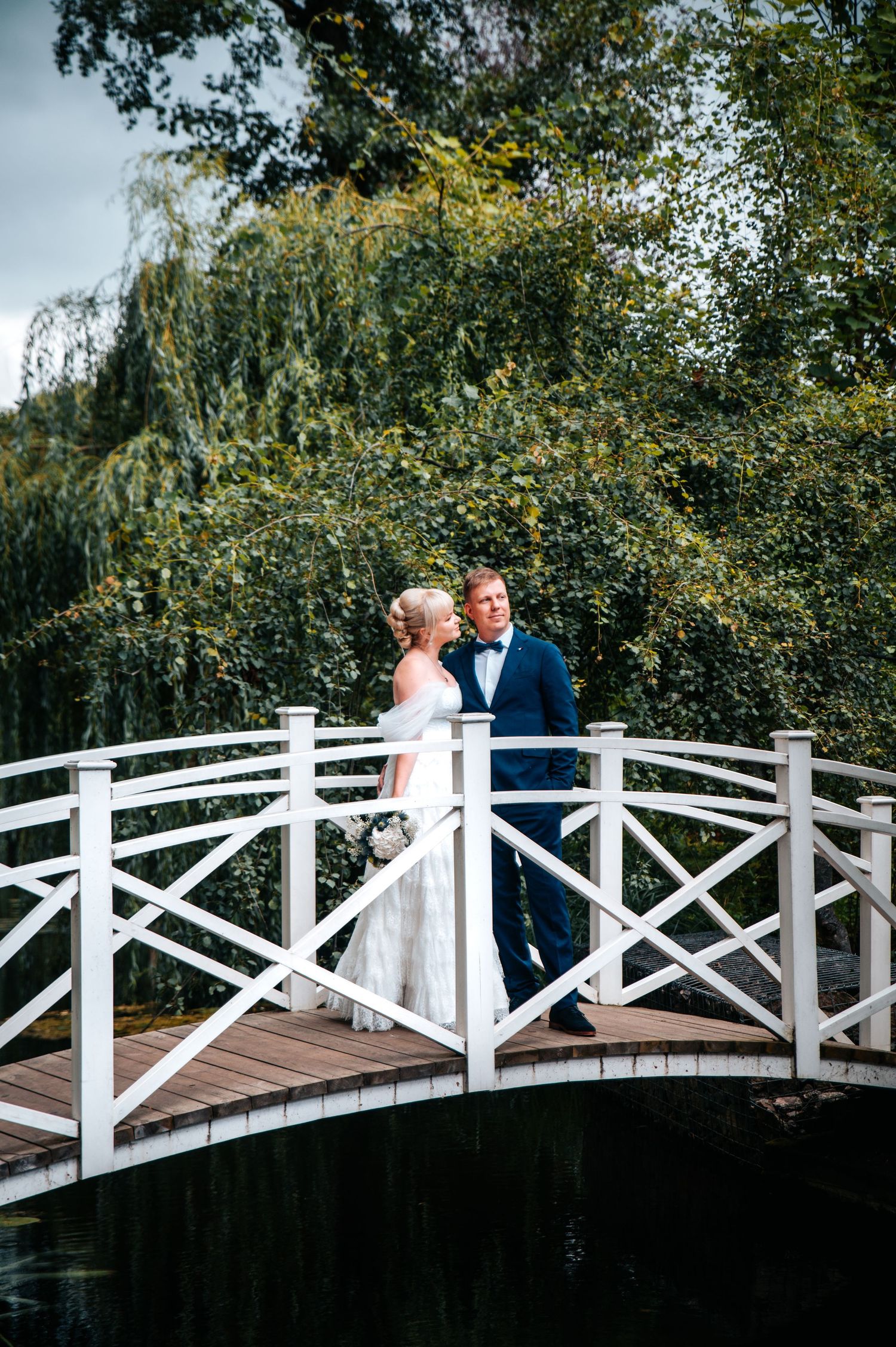 A couple shares a romantic moment on a white wooden bridge over water surrounded by lush green trees.
