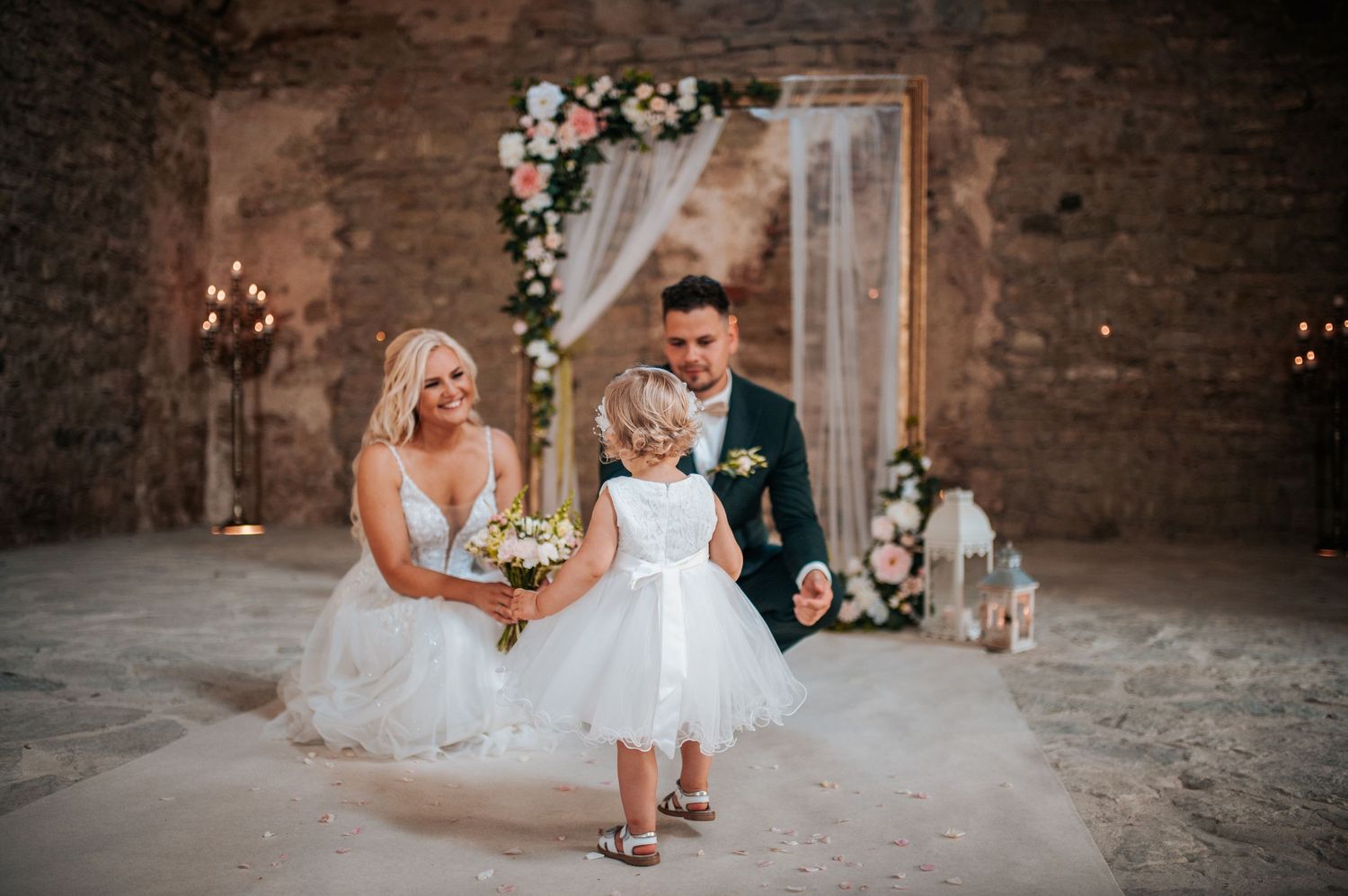 A wedding party walks through a rustic brick archway decorated with floral arrangements and string lights.