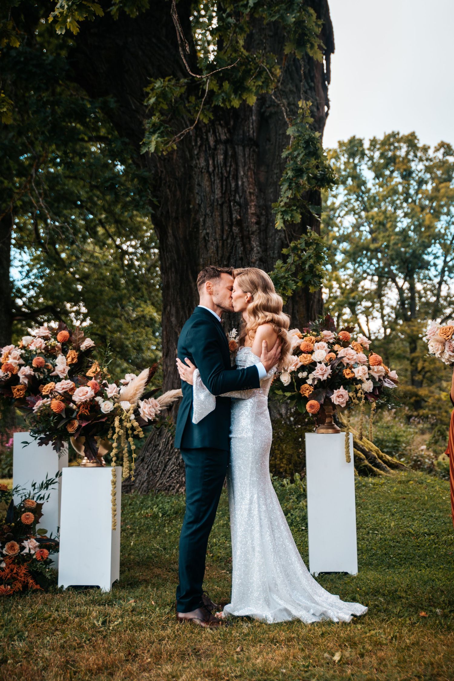 A romantic outdoor wedding ceremony setup with white pedestals and peach floral arrangements under a large tree.